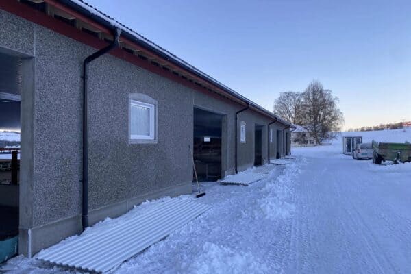 Nærbilde av landbruksbygning med isolert vegg, snødekt uteområde, landbruksmaskiner og ubebygde naturomgivelser i Innlandet, Norge.