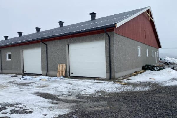 Landsbrygsbygg for landbruk i Innlandet med installerte garasjedører og peisstokk på byggets side, omgitt av snø og grus; ferdigstillelse av landbruksbygg i Innlandet.