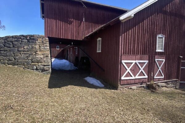 Gammel låvebygning i rød tre med hvite detaljer under en klar blå himmel, omgitt av jord og steinmur.