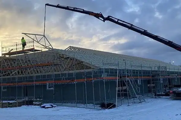 Takstolbygg under oppføring i kaldt vær, utendørs bygging av landbruksbygninger i Innlandet. Stillas og kran er i bruk for å sette opp takstrukturen. Landskap med snø og en skyet himmel.